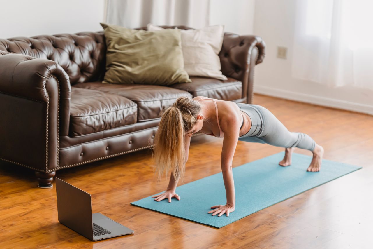 About Side view full body of sportive female in activewear practicing kumbhakasana asana on mat near laptop during online lesson in living room near couch