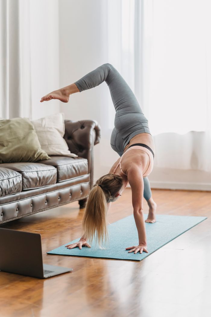 Full body of faceless female in sportswear performing eka pada svanasana posture while doing yoga near couch in living room with laptop