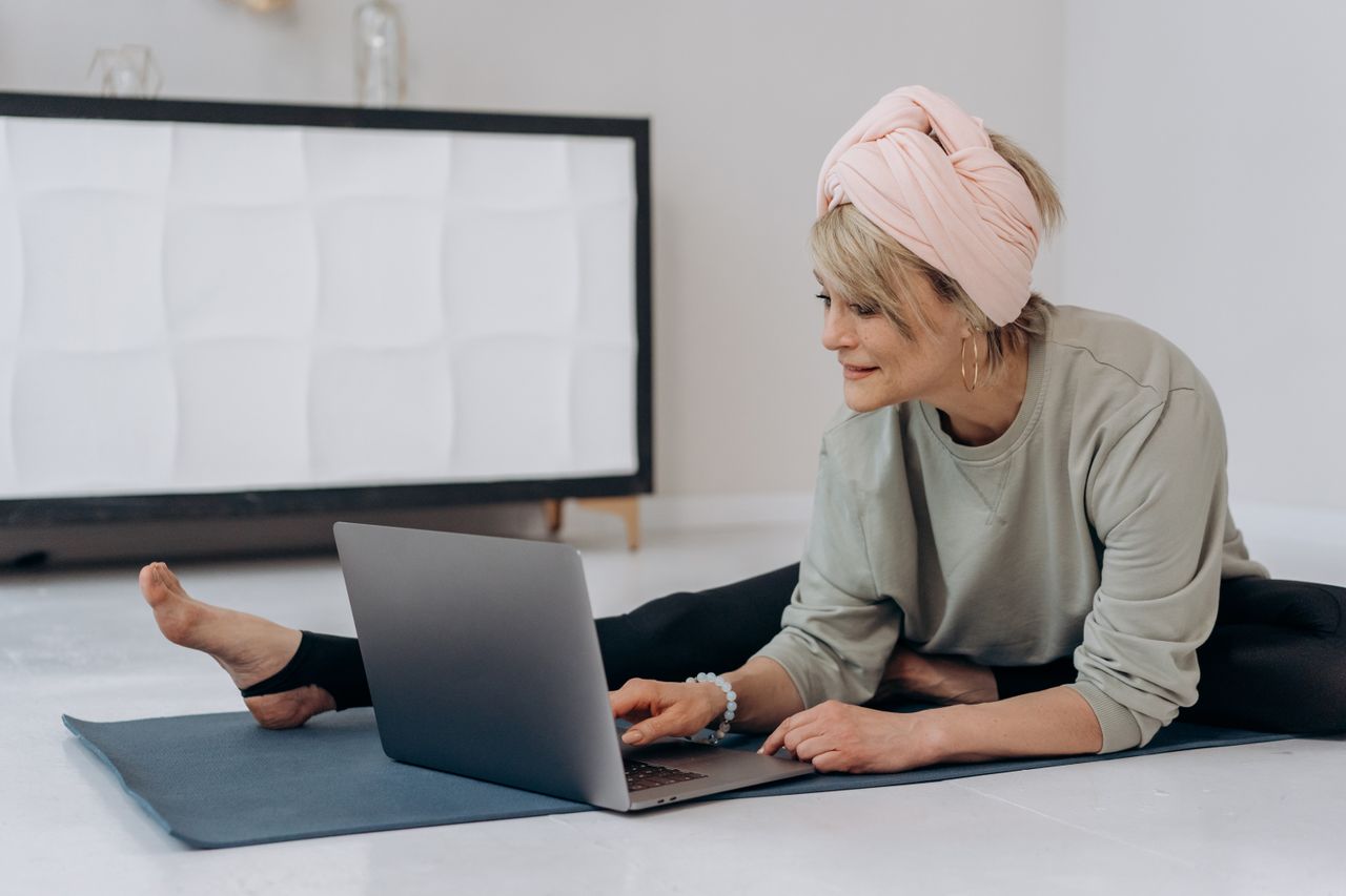 Contact A senior woman using a laptop while practicing yoga on a mat indoors, emphasizing active lifestyle.